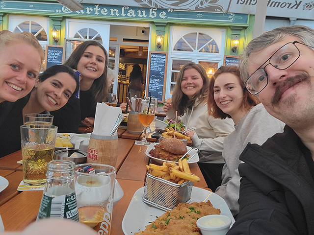 A group of six friends sitting at a restaurant table on a patio and posing for selfie