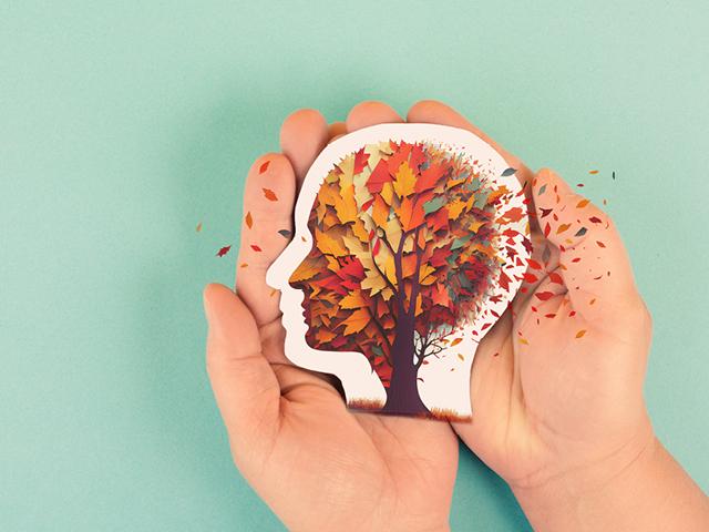 Two hands are holding an image of a tree in the shape of a human head; the leaves are autumanl colors and are begining to blow away